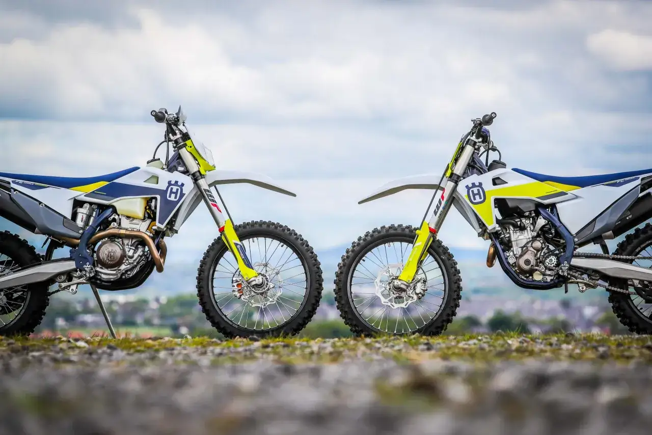 Due moto Husqvarna bianche e blu, pronte per l'enduro motocross, posizionate fianco a fianco su un terreno erboso con cielo nuvoloso.