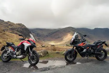 Due moto, una Ducati Multistrada 1290 rossa e bianca e una KTM 1290 Super Adventure nera e arancione, parcheggiate su una strada di montagna con un paesaggio nebbioso sullo sfondo.
